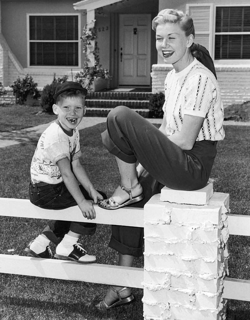 Image Credits: Getty Images / Hulton Archive | American actor and singer Doris Day smiles and laughs as she sits on a fence in front of a house with her son, Terry. Day wears sandals, trousers and has her hair tied back in a scarf.