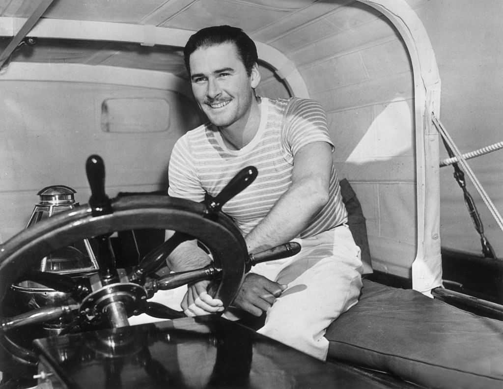 Image Source: Getty Images / Australian-born actor Errol Flynn (1909-1959) smiles as he sits with one hand on the captain's wheel of a yacht.