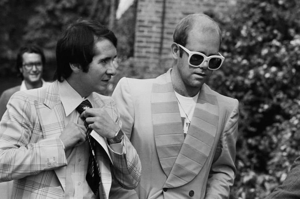 Image Credit: Getty Images / English singer, songwriter, pianist, and composer Elton John and his manager John Reid on their way to a luncheon in honour of Buddy Holly at the Orangery in Holland Park.
