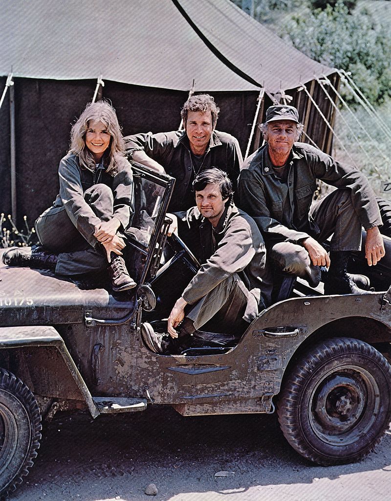Image Credits: Getty Images / Keystone | Alan Alda in the driving seat of a jeep, surrounded by Loretta Swit and other cast members of M.A.S.H