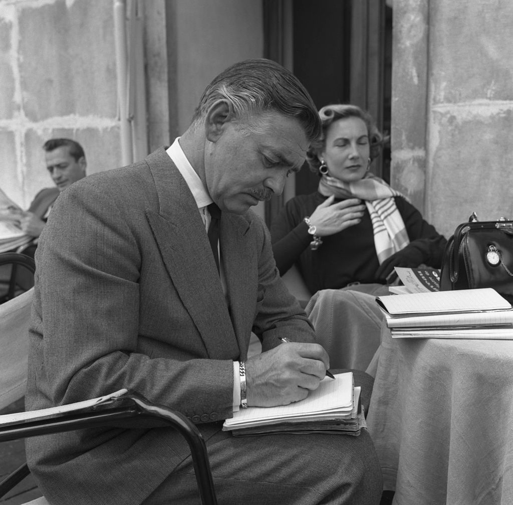 Image Credit: Getty Images / American actor Clark Gable writing next to his bride-to-be Kay Williams Spreckles sitting behind him in Venice, 1953. Image Credit: Getty Images / American actor Clark Gable writing next to his bride-to-be Kay Williams Spreckles sitting behind him in Venice, 1953.