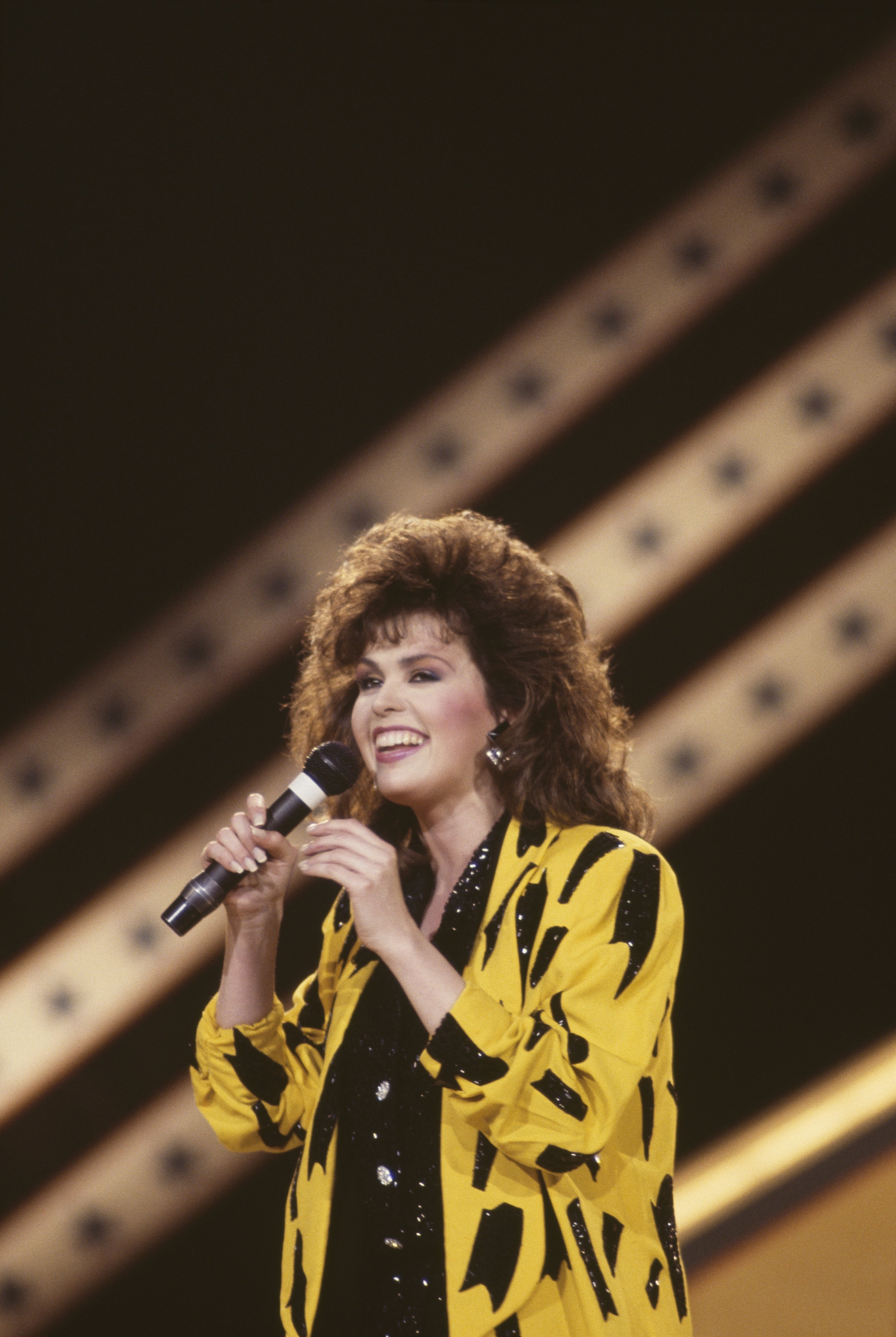 Image Credits: Getty Images / David Redfern / Redferns | Marie Osmond, US singer and musician, singing into a microphone during a live concert performance, on stage at the International Festival of Country Music, held at Wembley Arena, London, England, Great Britain, 1 April 1986.