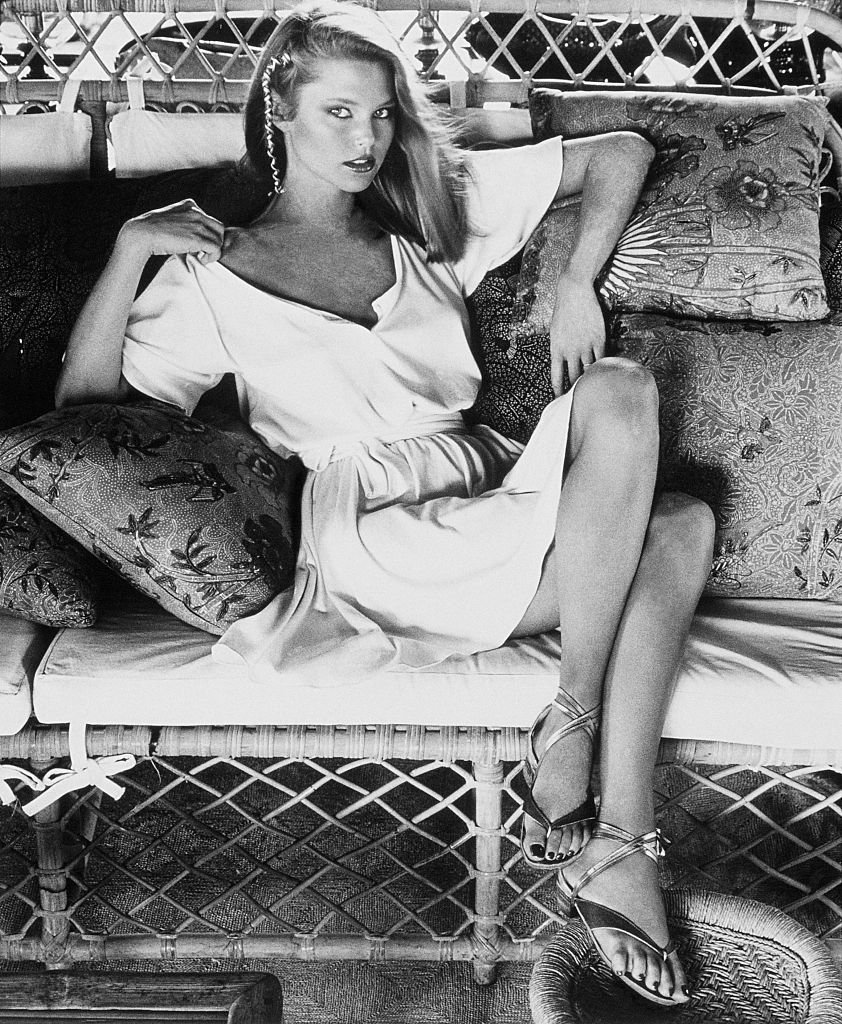 Image Credits: Getty Images / Kourken Pakchanian / Condé Nast | Model Christie Brinkley, sitting with crossed legs on a wicker couch with pillows, and wearing a short ivory T-shirt dress by Callaghan. Image Credits: Getty Images / Kourken Pakchanian / Condé Nast | Model Christie Brinkley, sitting with crossed legs on a wicker couch with pillows, and wearing a short ivory T-shirt dress by Callaghan.