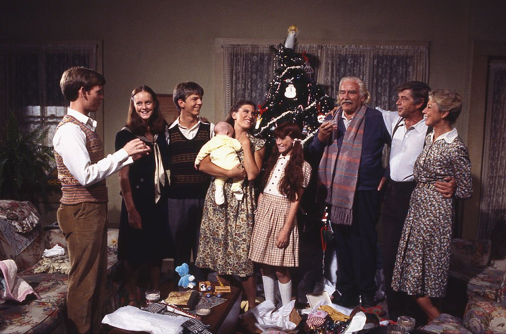 Image Credits: Getty Images / CBS | 'The Waltons' At Christmastime. American actors (left to right) Eric Scott as Ben Walton, Mary McDonough as Erin Walton, David Harper as Jim-Bob Walton, Judy Norton Taylor as Mary Ellen Walton, Kami Cotler as Elizabeth Walton, Will Geer (1902 - 1978) as Zeb Walton, Ralph Waite as John Walton, Michael Learned as Olivia Walton, gather to celebrate in front of a Christmas Tree during an episode of the long-running CBS Depression Era family drama series 'The Waltons,' California, 1977.