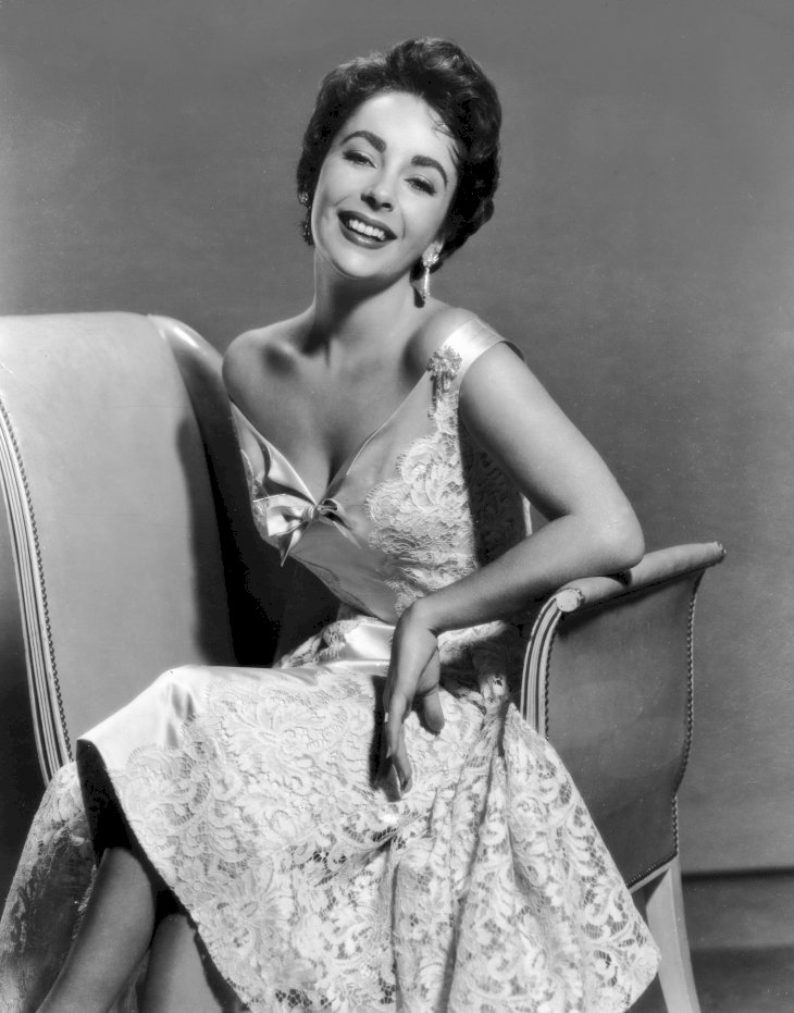 Image Credit: Getty Images/Hulton Archive | Elizabeth Taylor seated, wearing a sleeveless lace brocade dress.