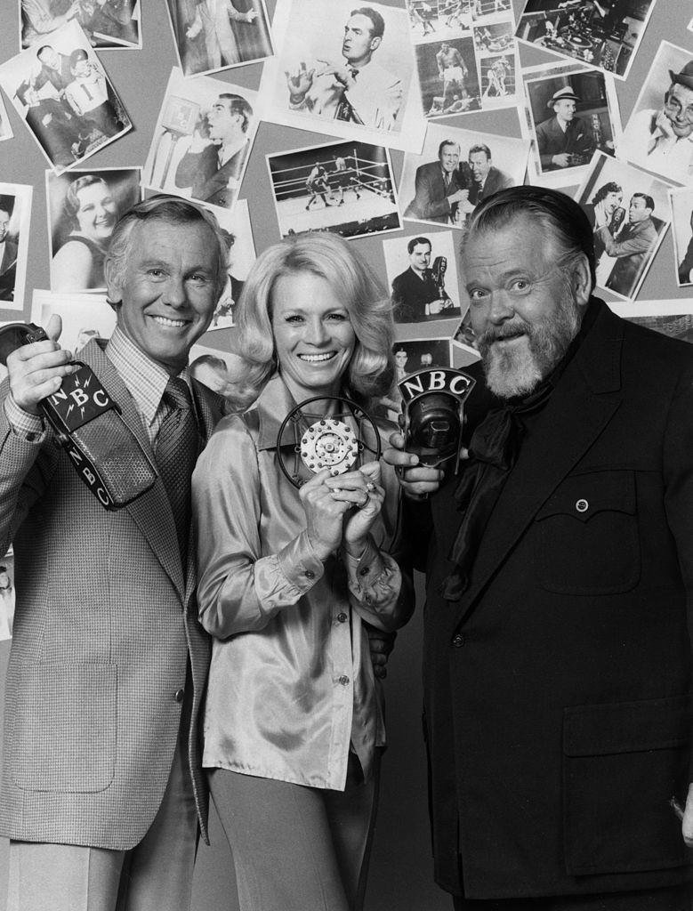 Image Credits: Getty Images / Hulton Archive | American television talk show host Johnny Carson, American actor Angie Dickinson and American film director and actor Orson Welles hold vintage NBC microphones and smile as they stand in front of a wall plastered with photographs of celebrities.