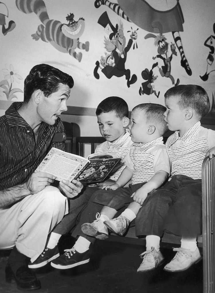 Image Credits: Getty Images / Hulton Archive | American television host Johnny Carson kneels while he reads a children's book to his sons (L-R): Kit, Cory and Ricky, in their nursery. There are cartoon characters on the wall behind them.