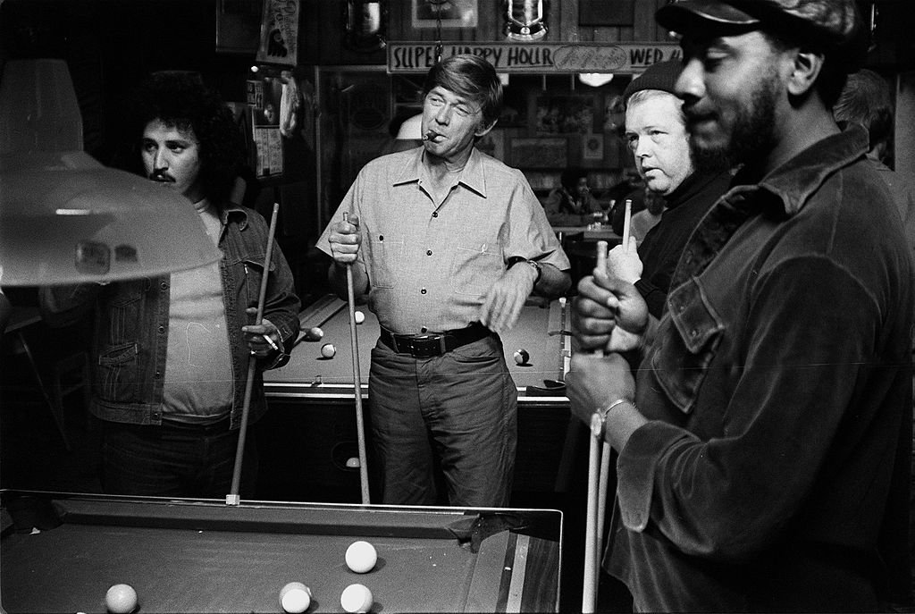 Image Credits: Getty Images / CBS | American actor Ralph Waite smokes a cigar and enjoys a game of pool with his buddies in a scene from the made-for-television movie 'The Secret Life of John Chapman,' 1976. Waite stars as college president John Chapman who goes on sabbatical and takes a series of blue-collar jobs in order to see what life was like outside of the halls of academia in this GE Theater Special.